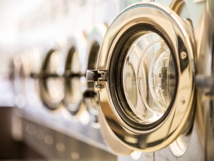  Close-up of a row of commercial washing machine doors in a laundromat, with the first chrome door open and the others receding in a blurred line.